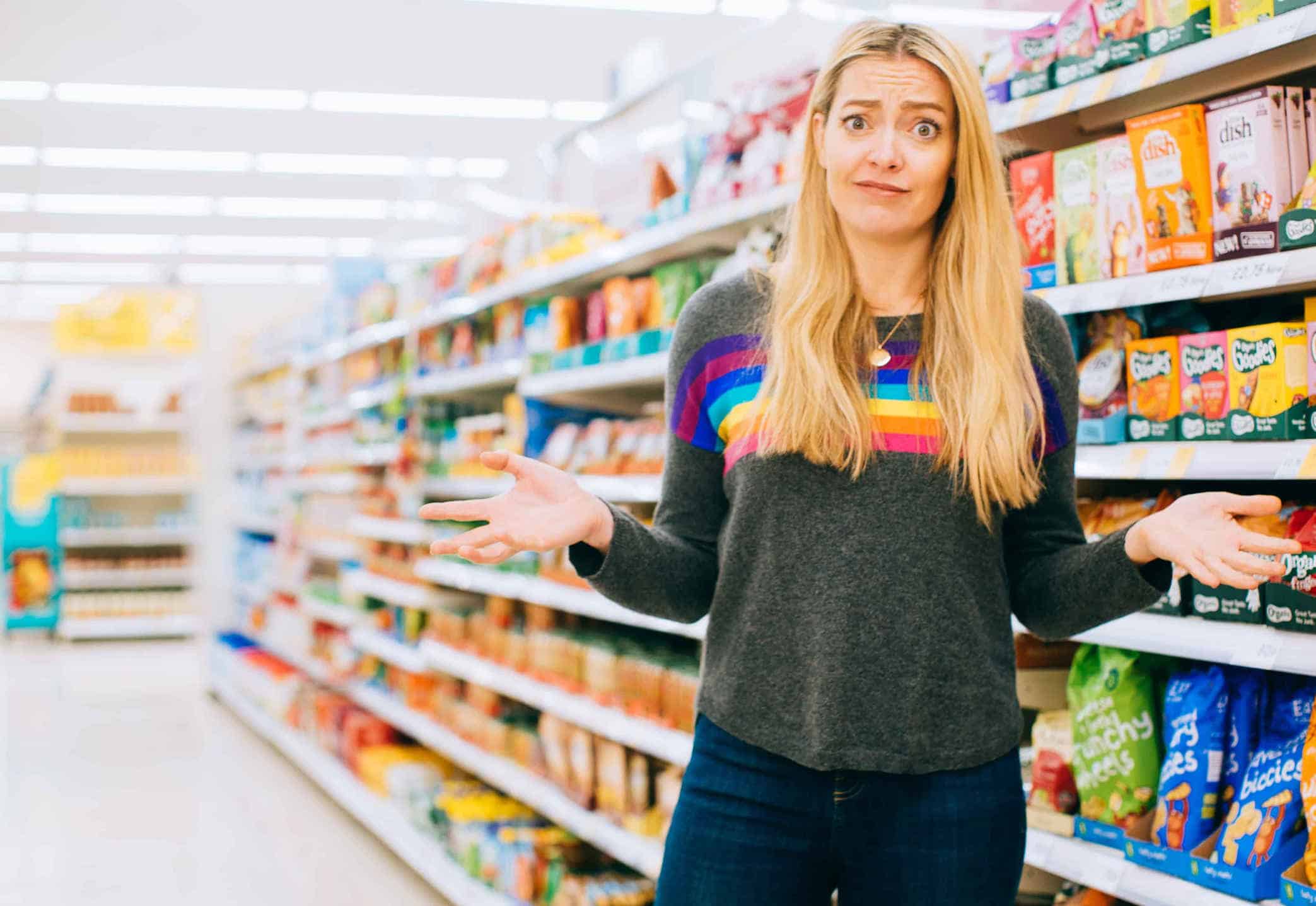 White person with long blonde hair stands in front of a supermarket aisle with a puzzled look on their face.