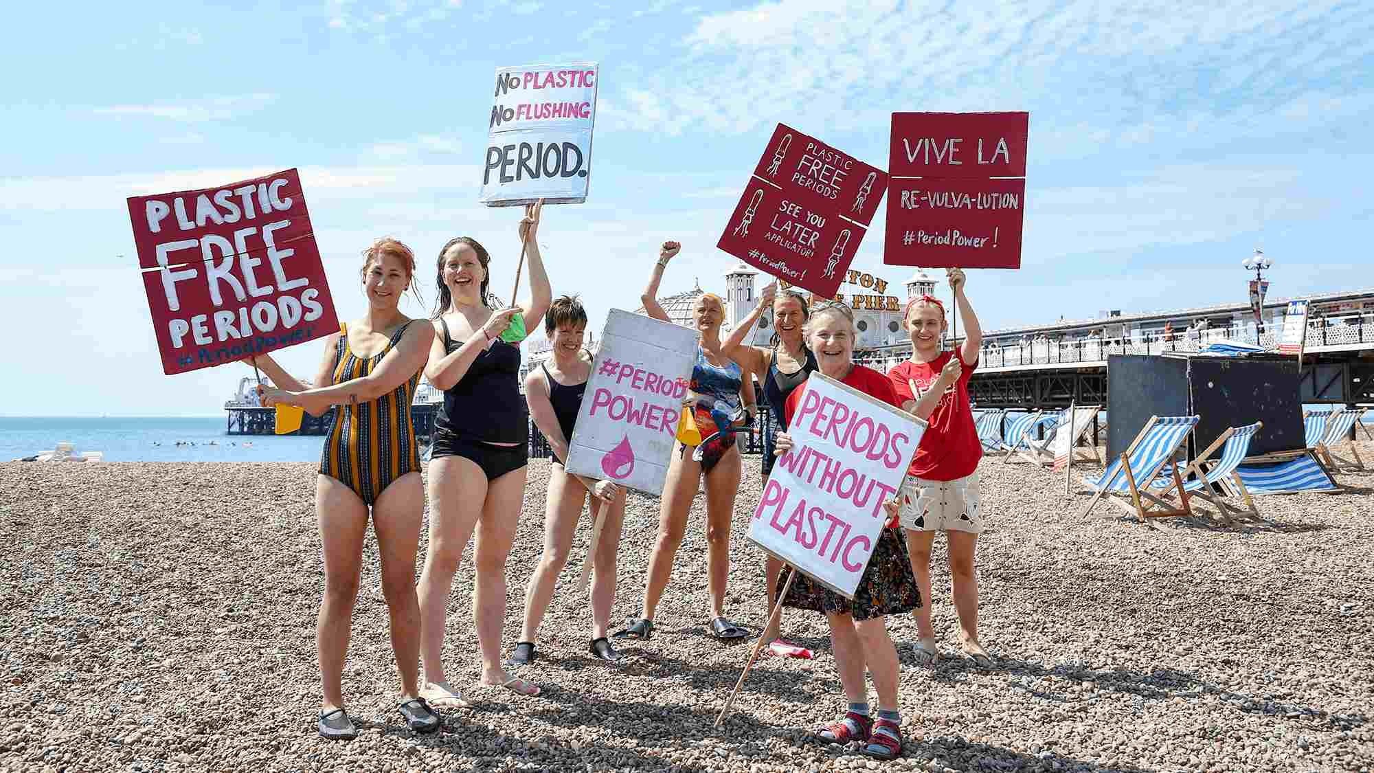 Seven women in swimsuits on a pebble beach holding placards that say "plastic free periods".