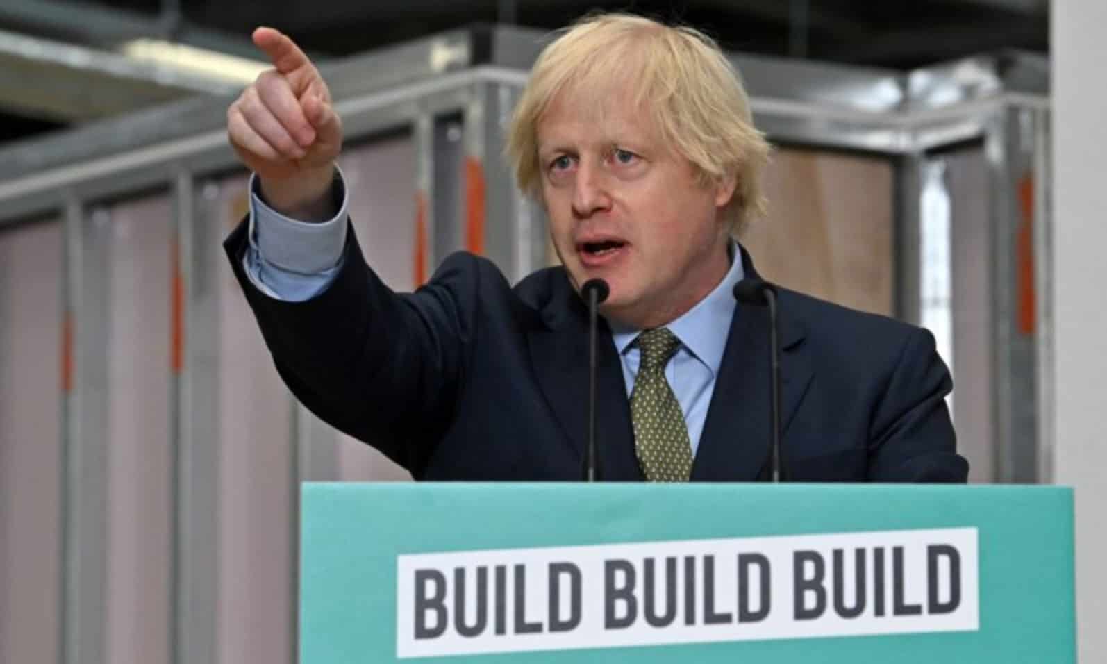 A white man in a suit points his finger, and speaks from behind a stand which has 'Build Build Build' written on the front