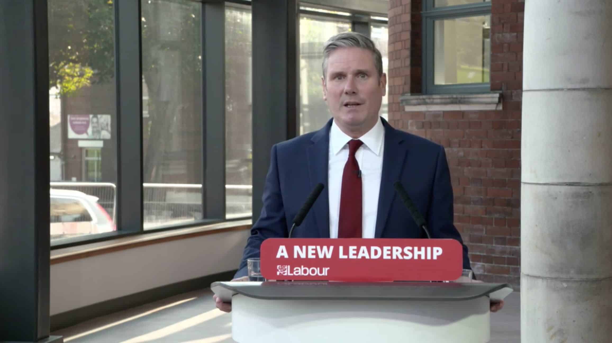 A white man in a suit stood at a press stand where a sign reads ' A New Leadership / Labour'