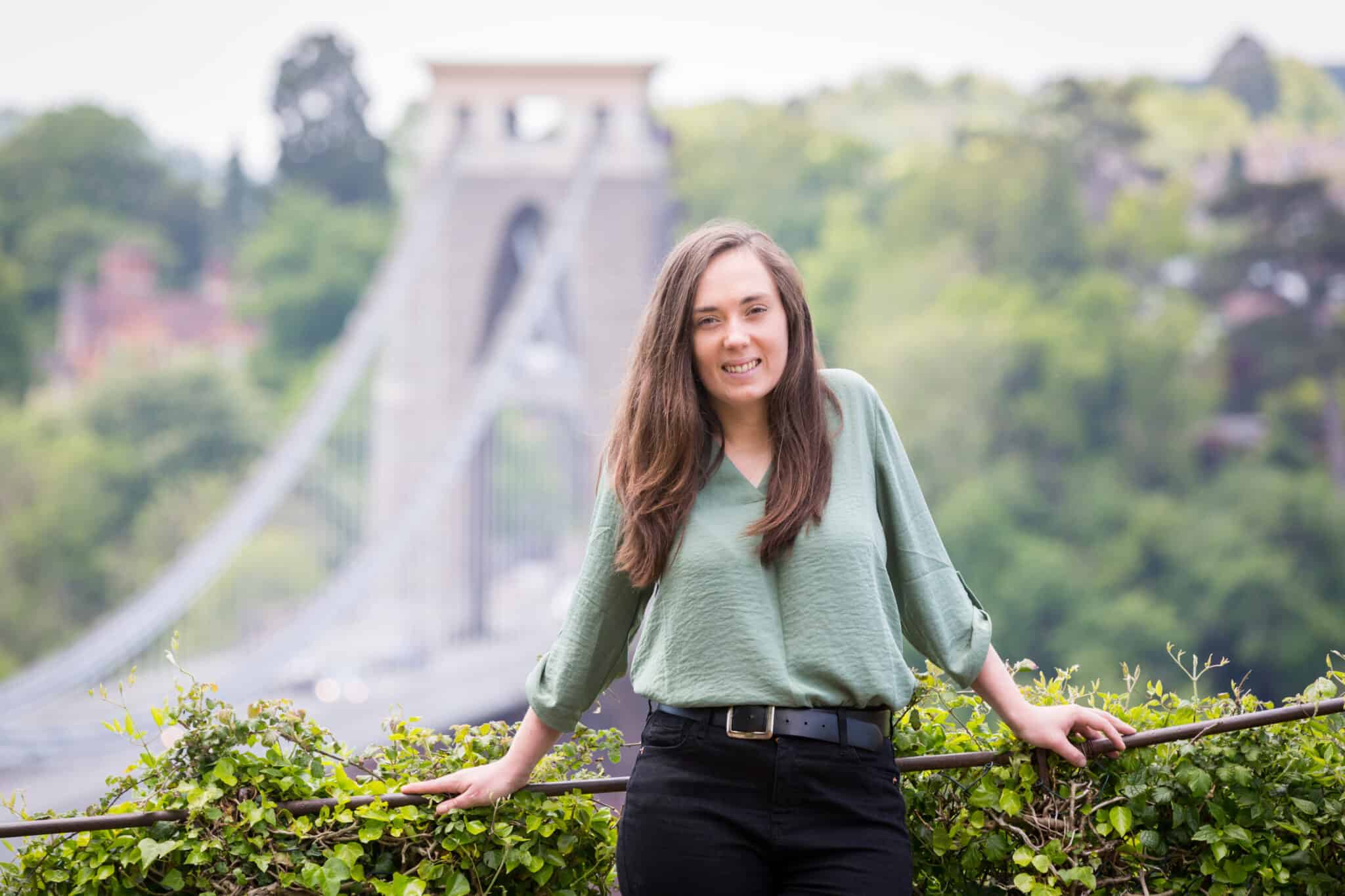 Jessie is leaning on a railing in front of Clifton Suspension Bridge. She is wearing a green blouse and black jeans.