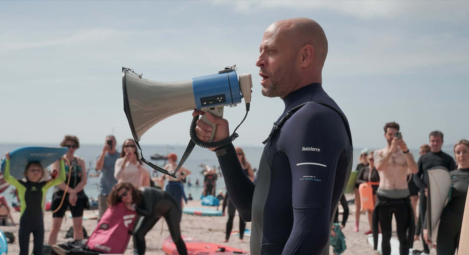 Hugo Tahholm, CEO of SAS, is pictured from the waist up in a wetsuit. He holds a megaphone and speaks through it to assembled protestors on the beach, who can be seen in the background.