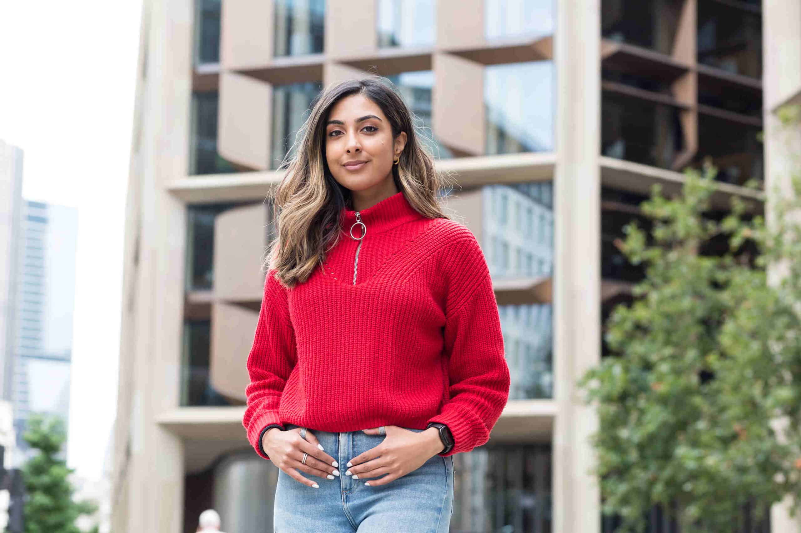 Kiran has brown hair and is stood in front of a tall building. She wears light blue jeans a red quarter-zip jumper and has her hands in her pockets.