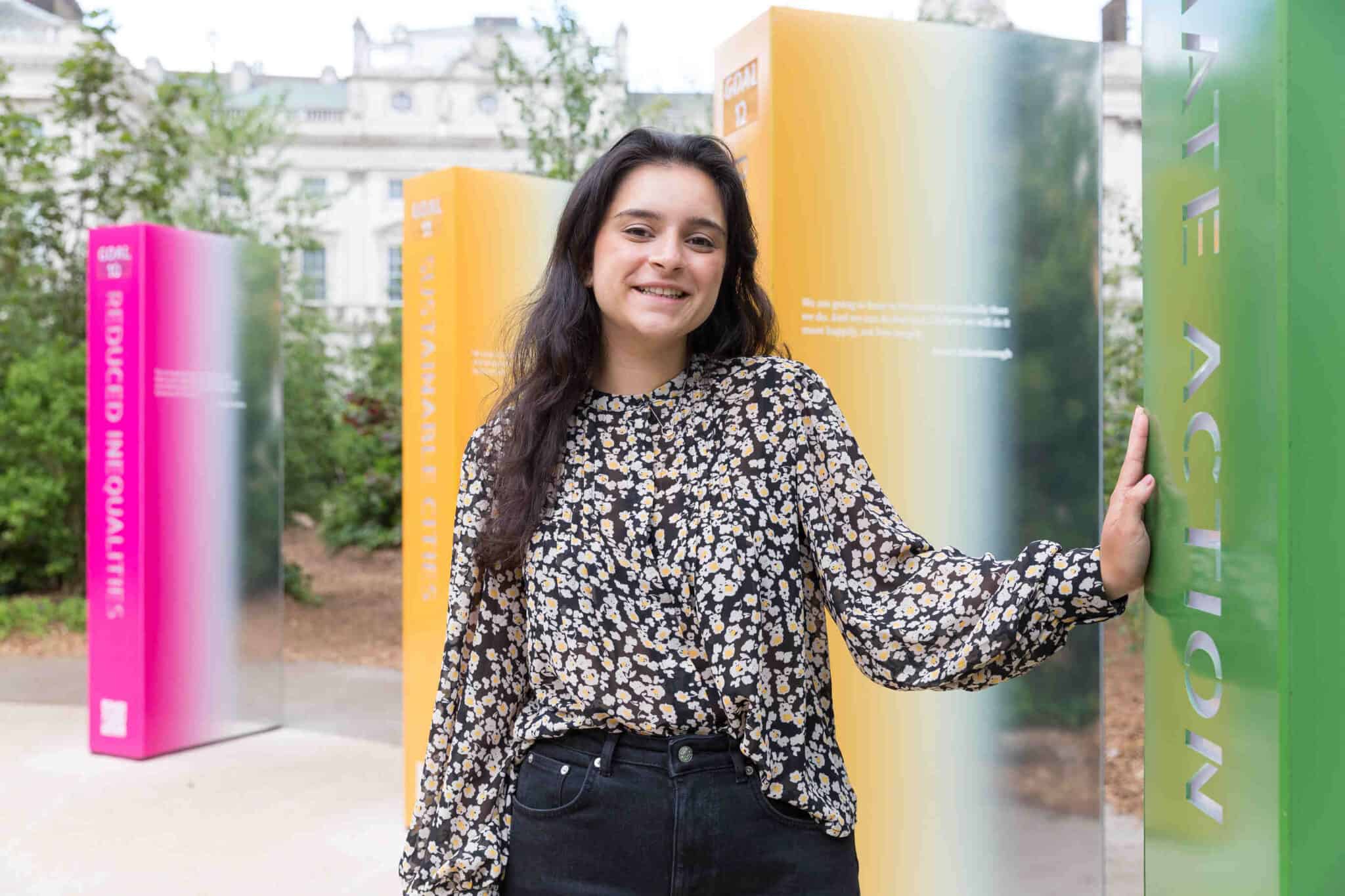 Sindhu has long black hair, and is standing in a cricle of pillars that represent the 17 sustainable development goals. She is wearing a floral print long sleeve blouse and black jeans.