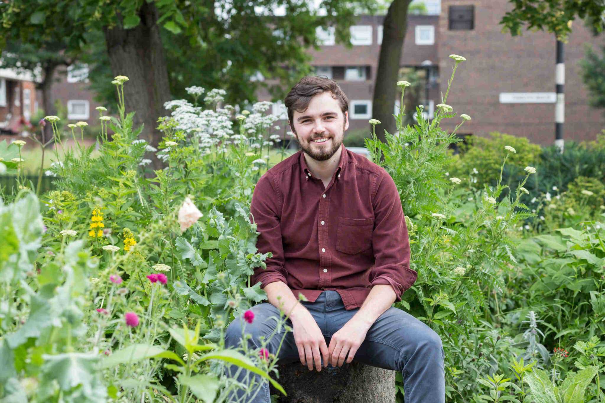 Joe is sat on a tree stump in the midst of a wildflower garden. In the distance is a block of red brick flats and a black and white sign post. Joe is wearing a red shirt and blue chinos.