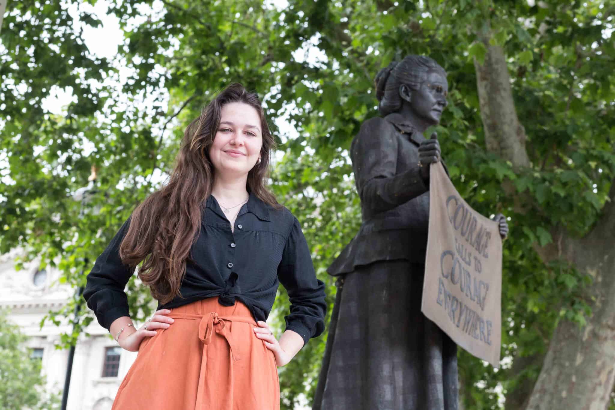 Madeleine is standing in front of Millicent Garrett Fawcett's statue in parliament Square. Her hands are on her hips.