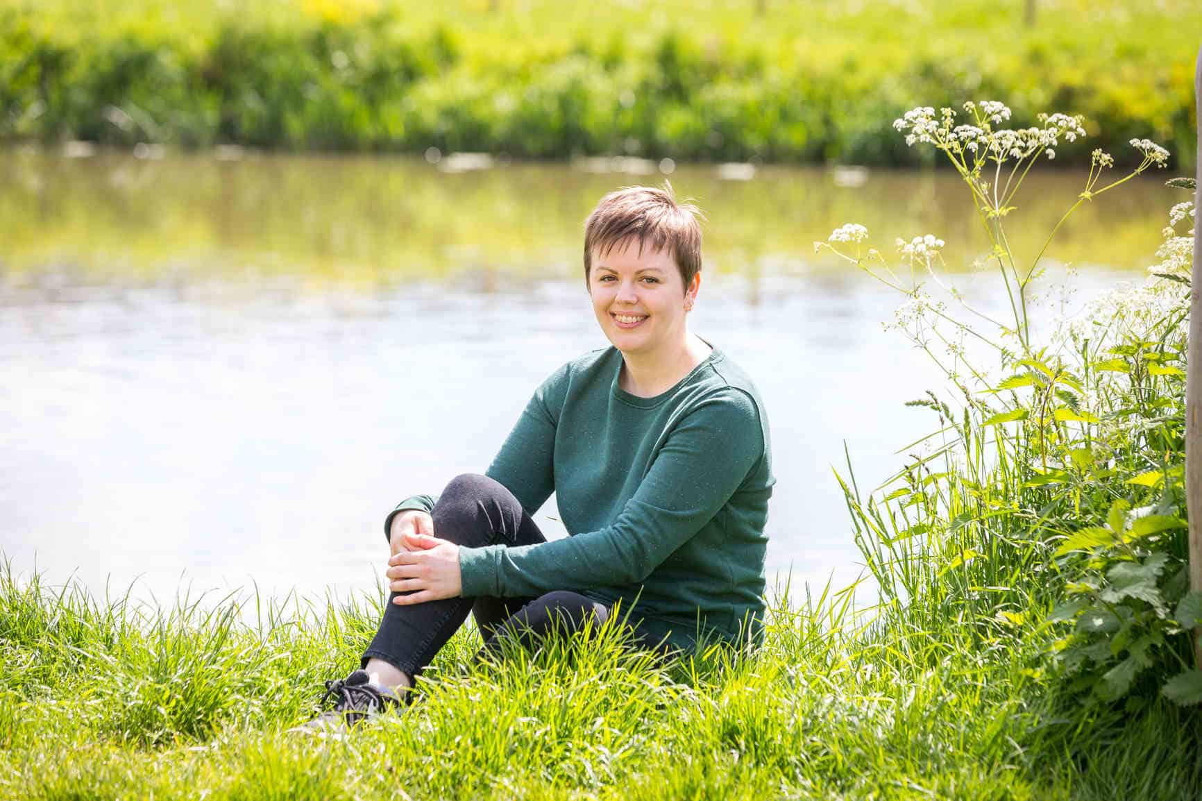 Fiona is sitting on the bright green grass in front of the River Avon. Behind her you can see the opposite riverbank. Fiona has one leg tucked into her chest which she is holding with her hands in a relaxed pose. She is smiling, wearing a green sweater, black skinny jeans and black trainers.