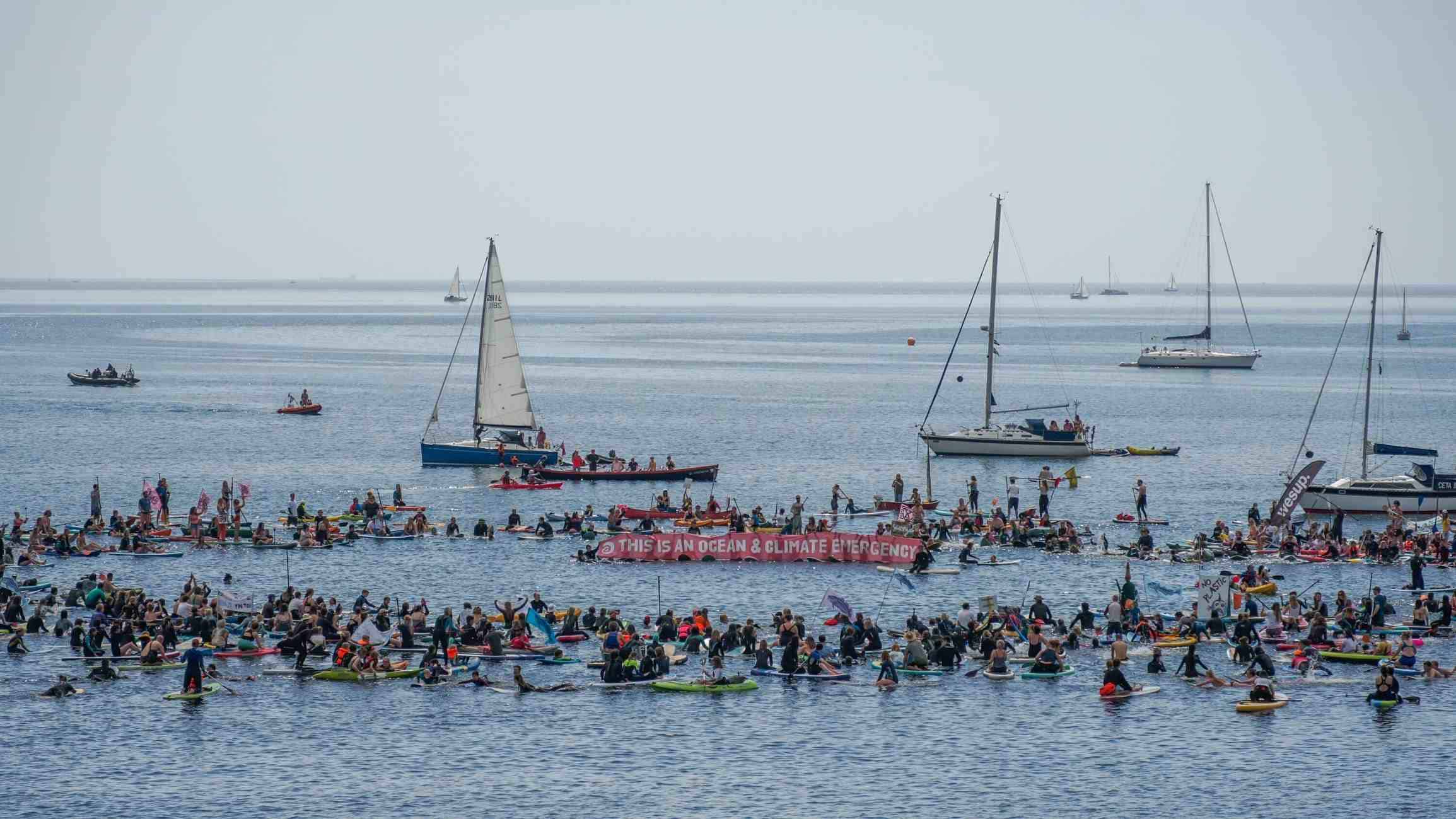 Wide-angle shot showing hundreds of people in a blue-grey sea. They have formed a ring, and in the centre of the ring protesters hold a long red banner that reads 'This is an ocean &climate emergency' alongside the SAS logo. Various yachts are dotted in the background.