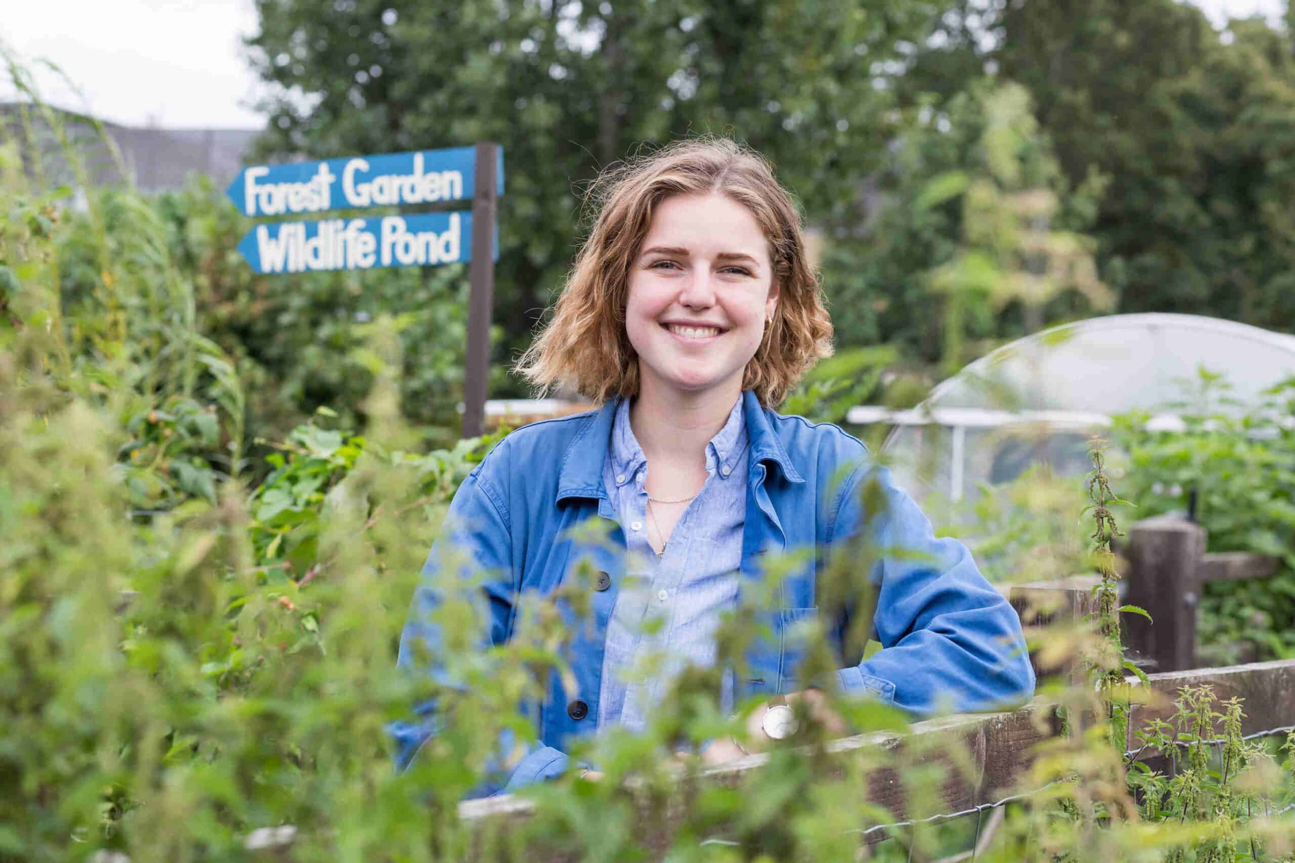 Georgia has light brown curly hair. She is wearing an electric blue chore jacket and light blue linen shirt. She's leaning on a fence surrounded by plants.