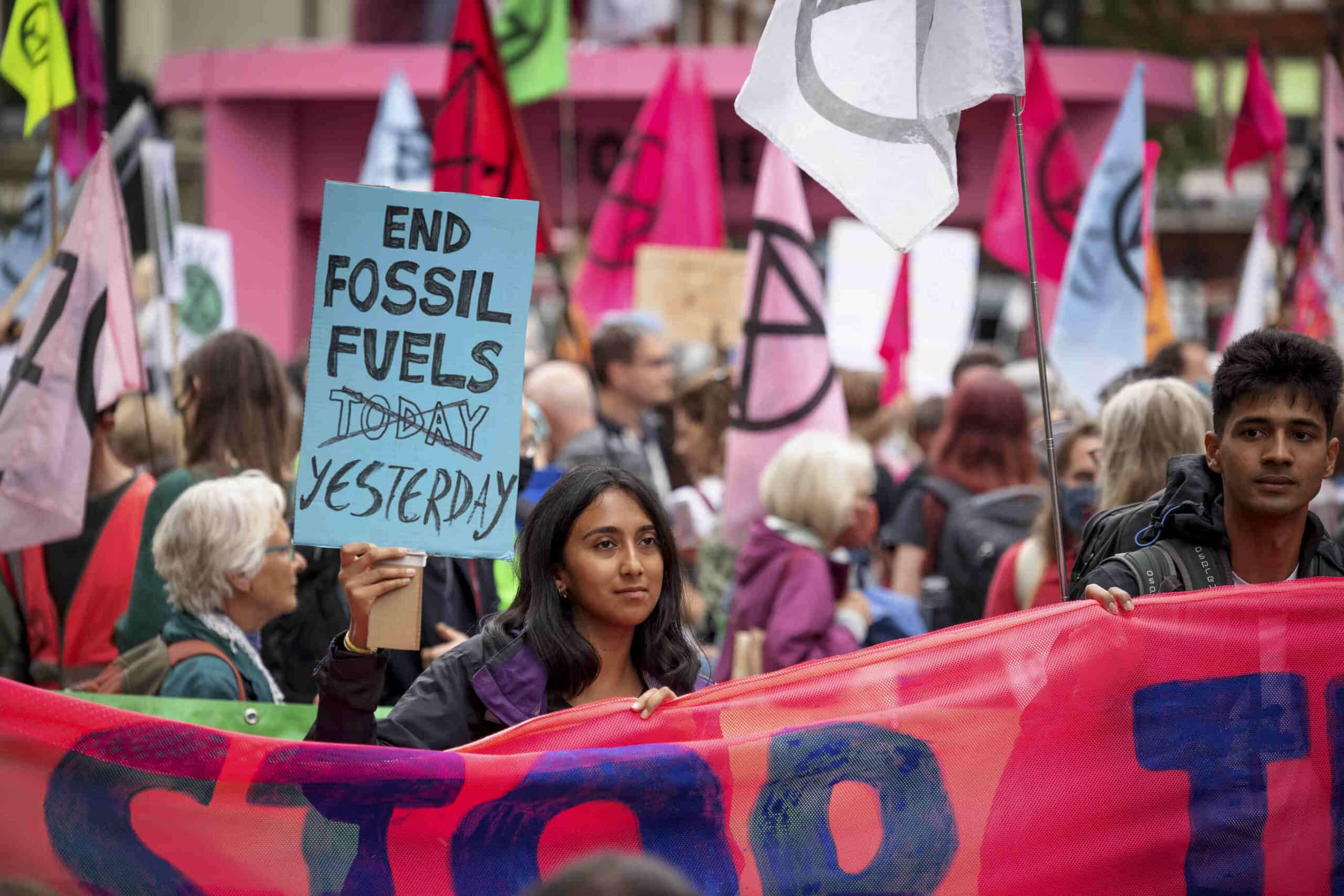 Two People of Colour are standing behind a large pink banner holding protests signs that say 'End Fossil Fuels yesterday". Behind them are many many protestors holding similar banners and placards.