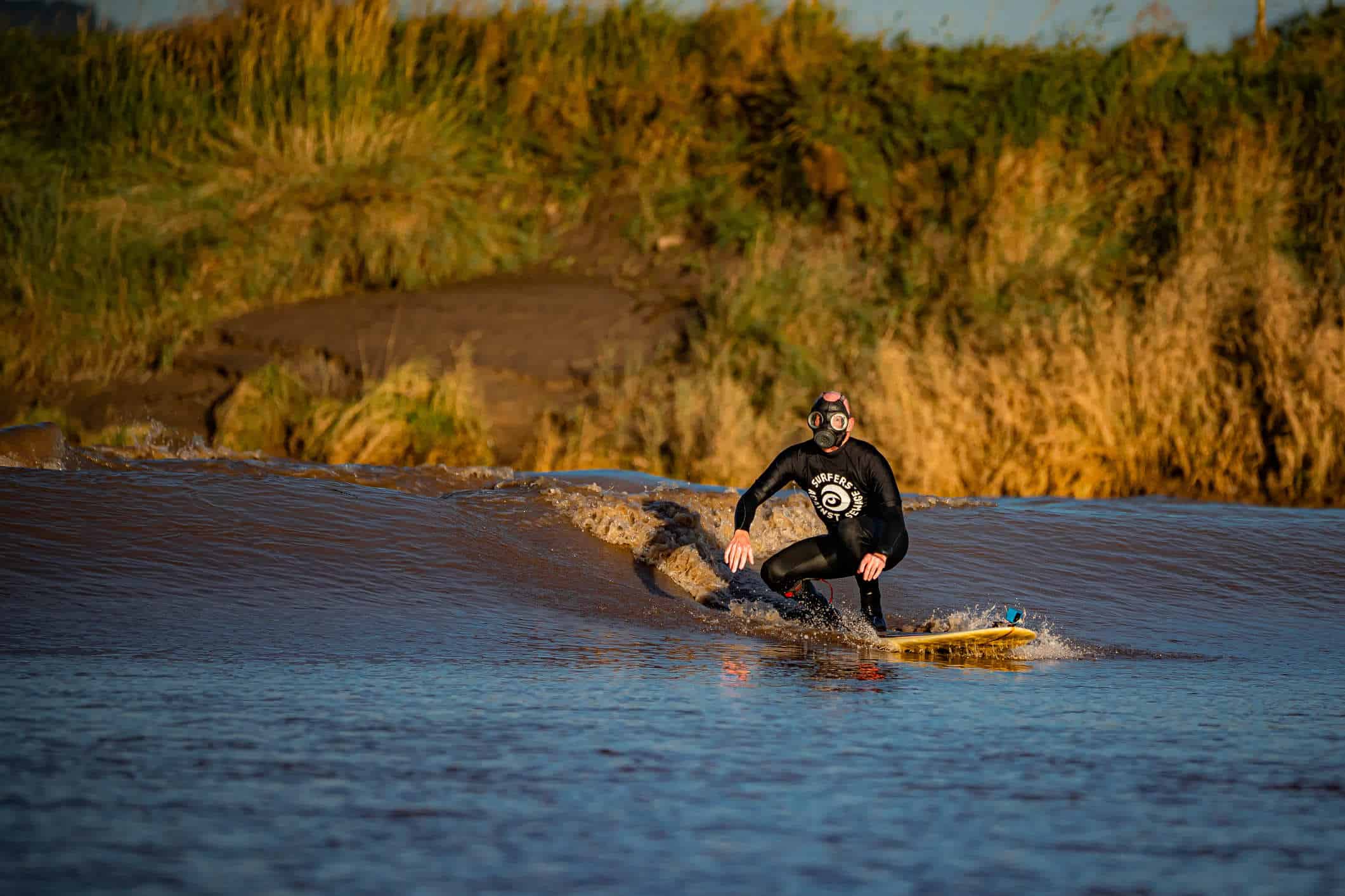 A person in a black wetsuit and wearing a black gas mask surfs a brown muddy wave.