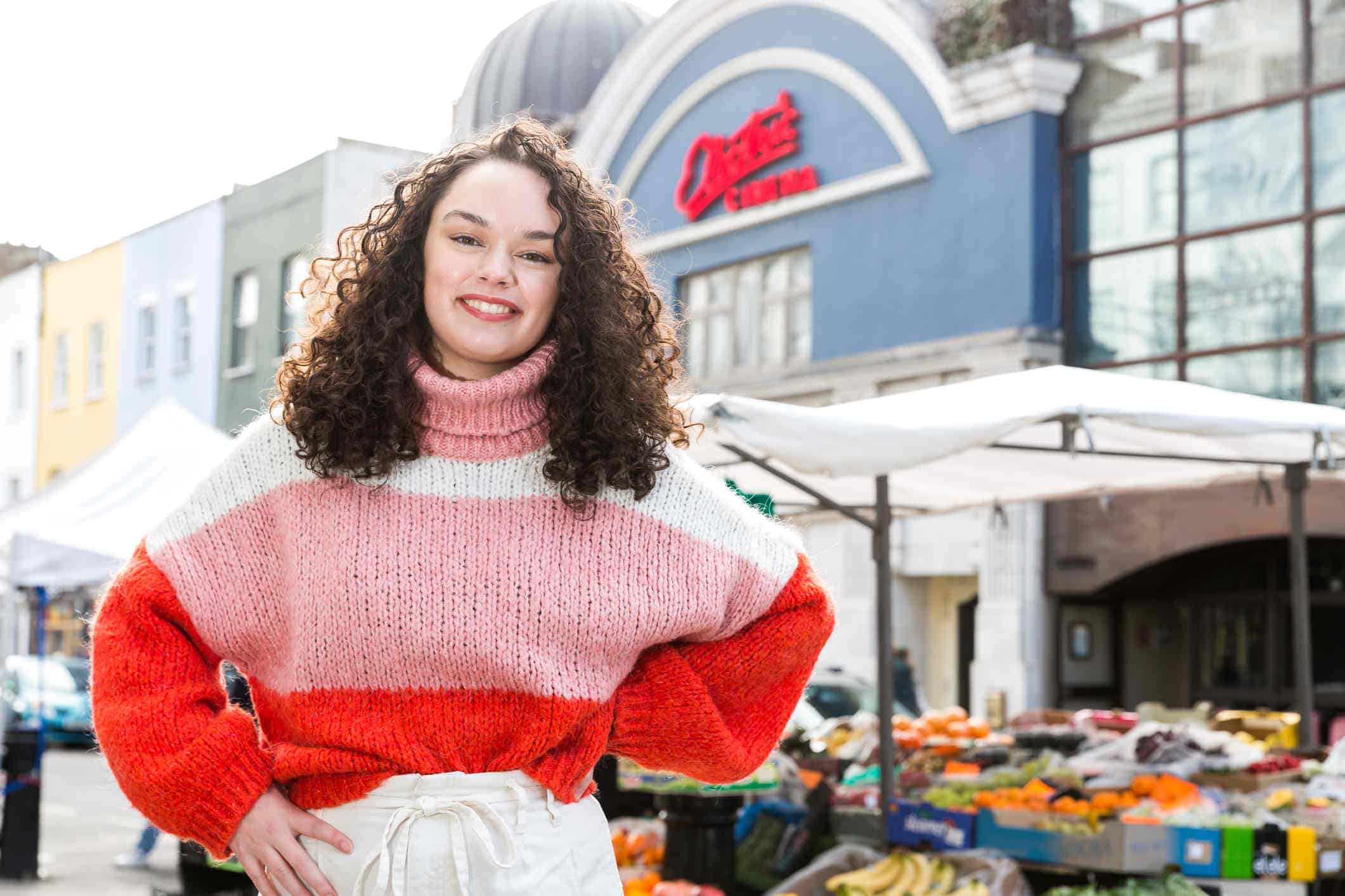 A woman is stood in front of a market with her hands on her hips, smiling. She has curly dark brown hair and is wearing a pink, red and white striped jumper.