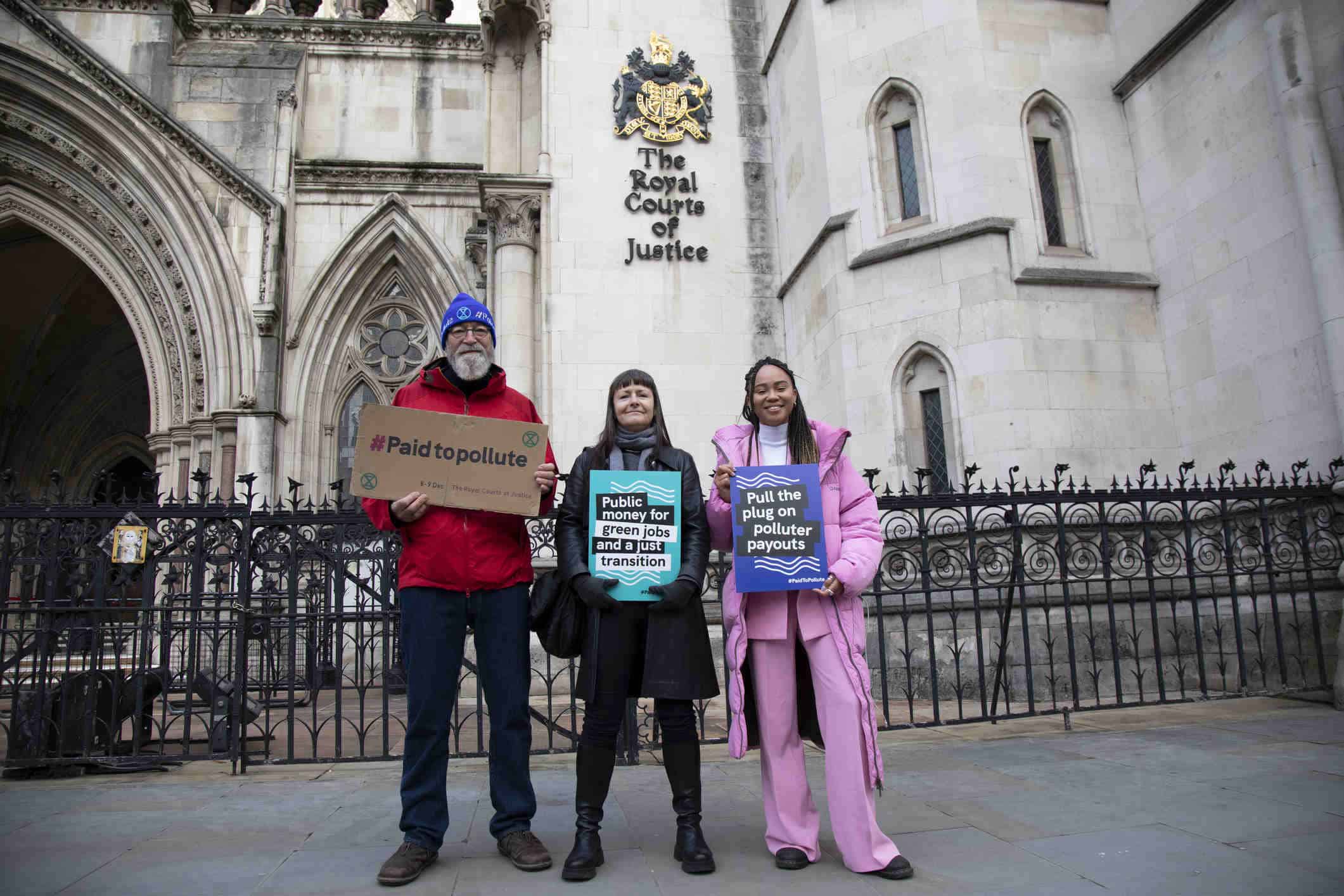 Three people stood outside the Royal Court of Justice holding placards that say "Paid to Pollute"