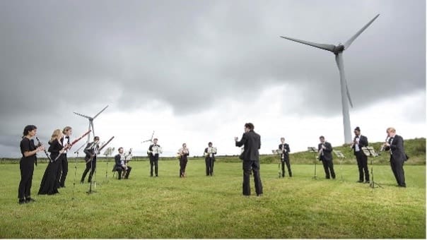 Musicians playing on a green field in front of a wind turbine.